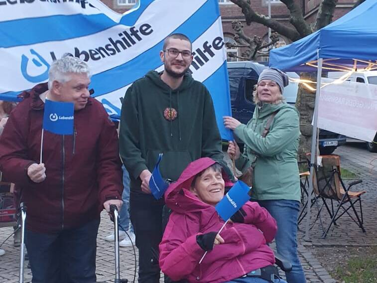 Drei Personen mit Lebenshilfe Flagge, eine davon im Rollstuhl.