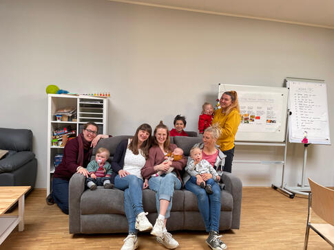 Mütter mit Ihren Kindern sitzen oder stehen an einem grauen Sofa