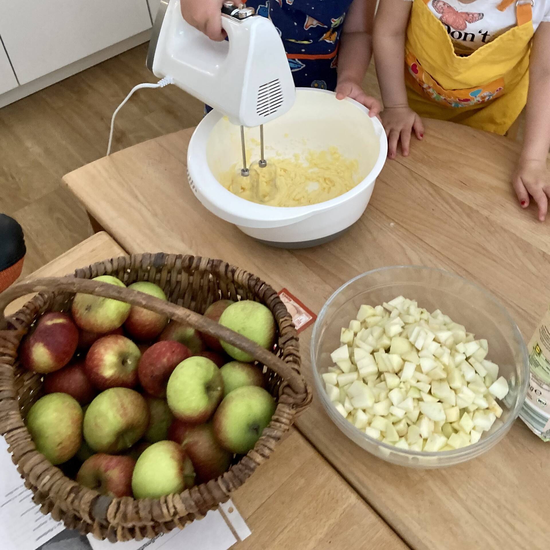 Zutaten für den Apfelkuchen und eine hand am Mixer der in einer Schüssel Teig rührt.