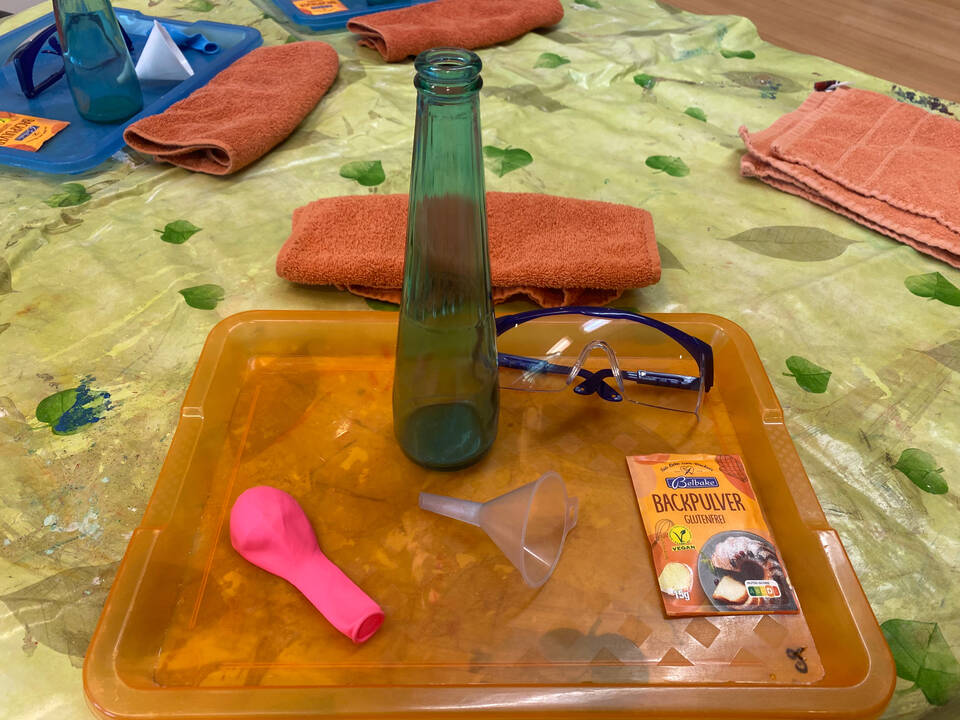 Ein Trichter, ein rosa Ballon, eine Schutzbrille und Backpulver liegen auf einem Tablett.