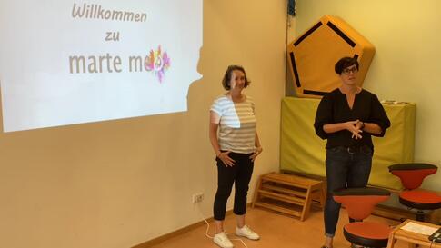 Zwei Frauen stehen in einem Raum vor einer Wand mit einer Präsentation.