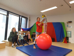 Ein Kind steht mit Gehörschutz auf einem roten Gymnastikball und wird von einer Frau festgehalten.