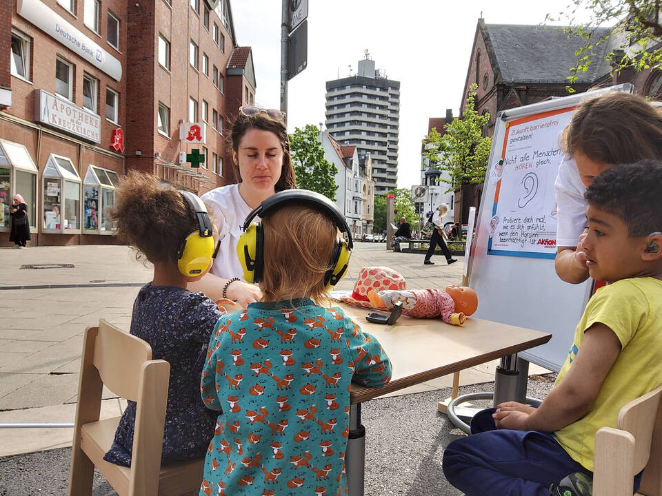 Kinder mit Schallschutzköpfhörereb stehen an einem Tisch bei einer Frau.