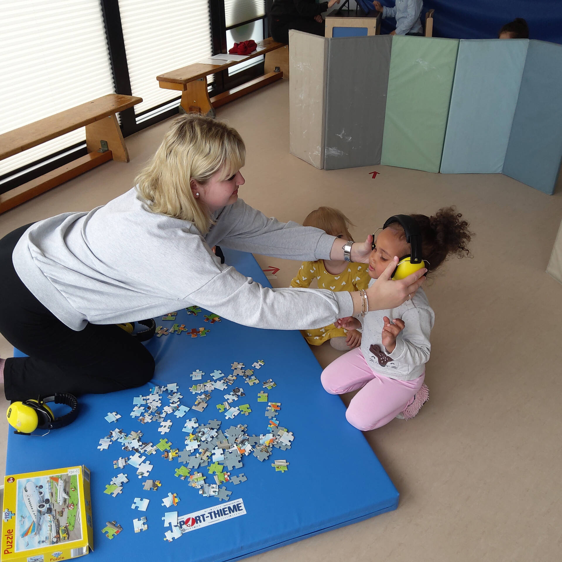 Eine Frau setzt einem Kind Kopfhörer auf, sie hocken auf dem Boden vor einem Puzzle.