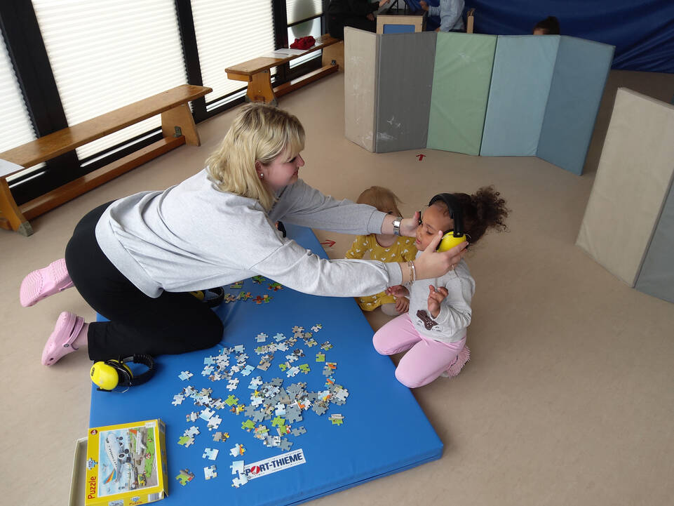 Eine Frau setzt einem Kind Kopfhörer auf, sie hocken auf dem Boden vor einem Puzzle.