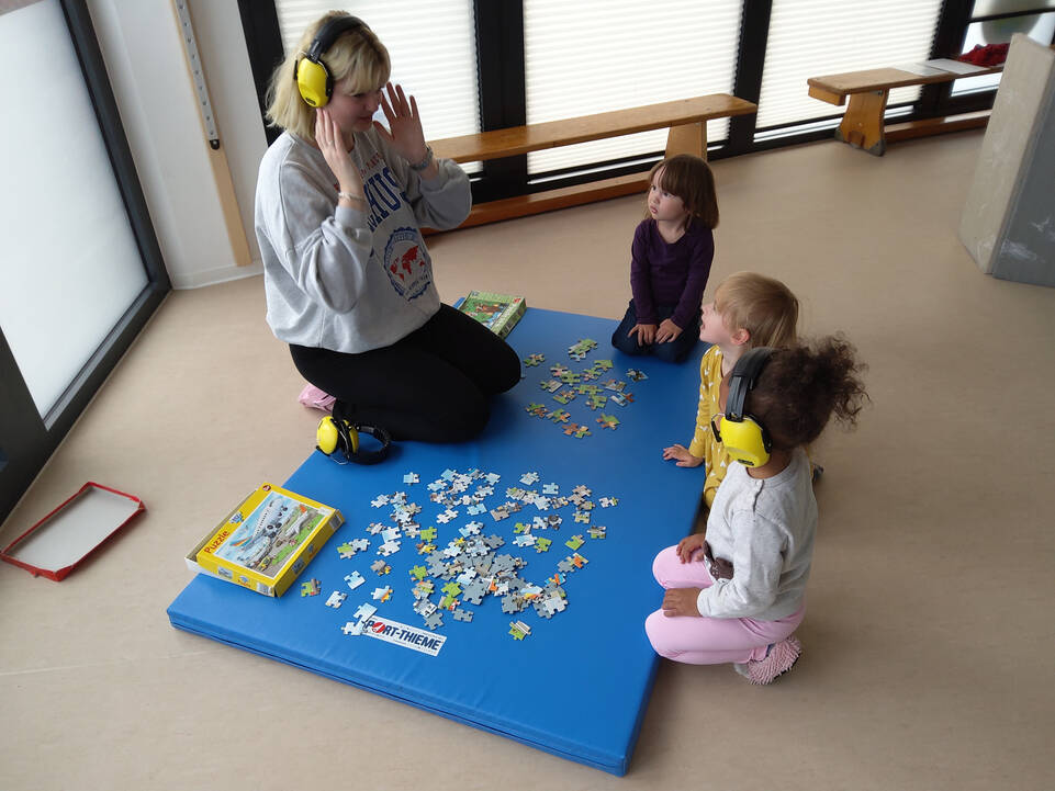 Eine Frau setzt einem Kind Kopfhörer auf, sie hocken auf dem Boden vor einem Puzzle.