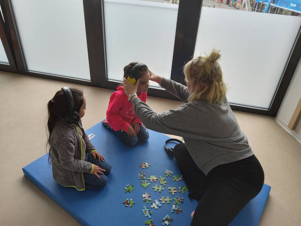 Zwei Kinder, eins hat Kopfhörer auf dem Kopf, das Andere bekommt gerade welche angezogen.