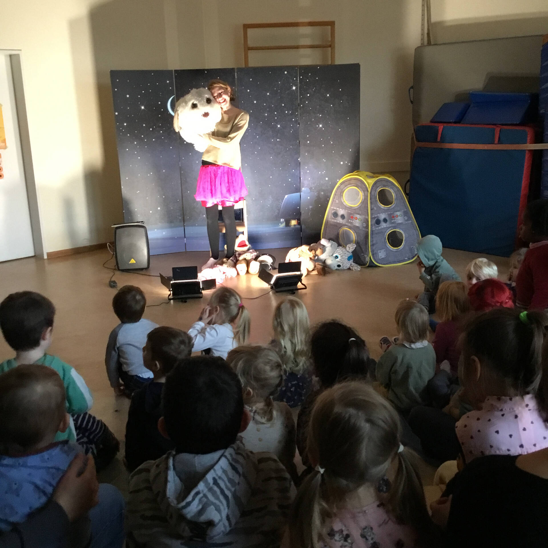 Einige Kinder sitzen auf dem Boden und schauen einer Frau vor einer Pappwand zu.