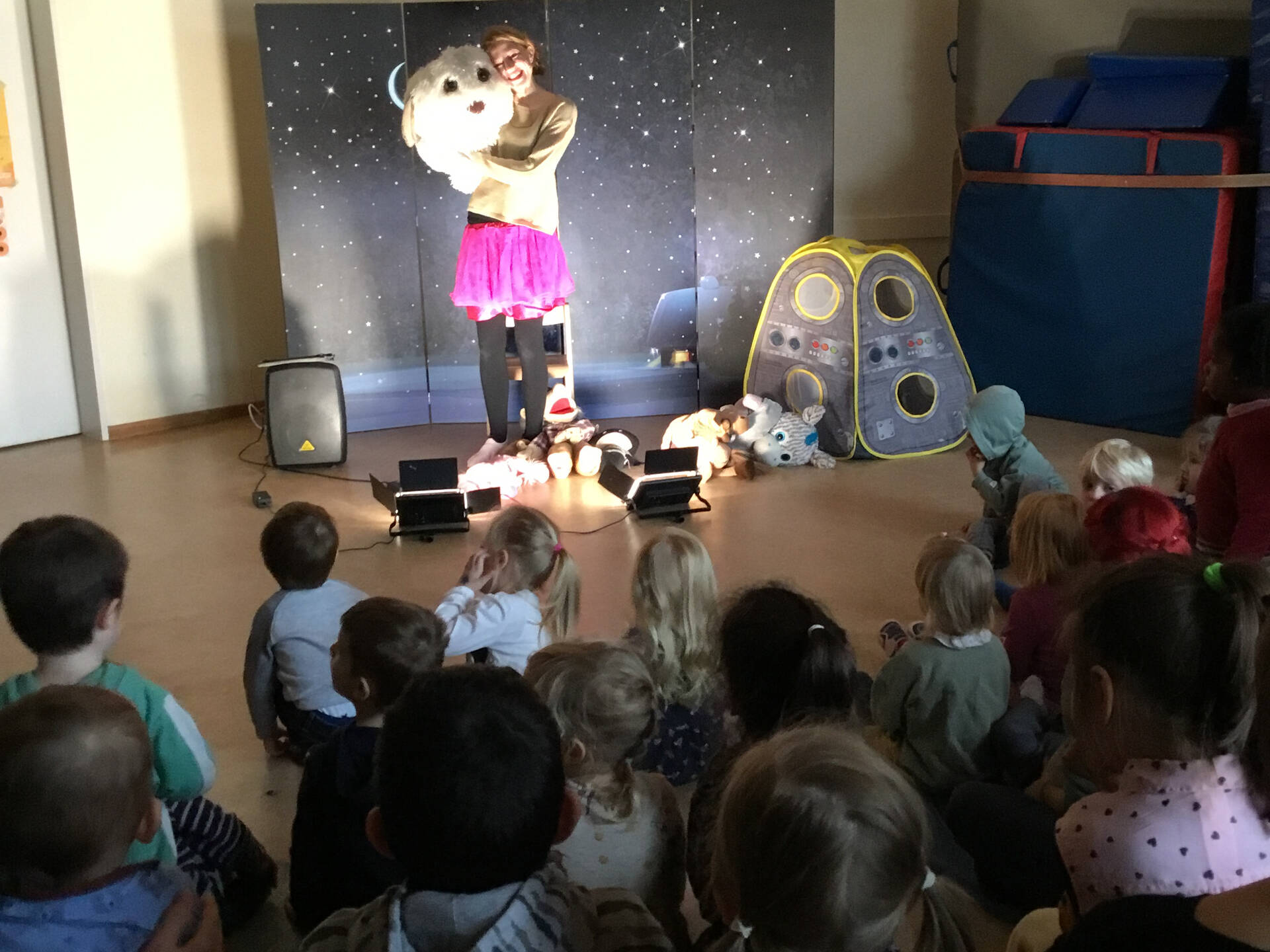 Einige Kinder sitzen auf dem Boden und schauen einer Frau vor einer Pappwand zu.