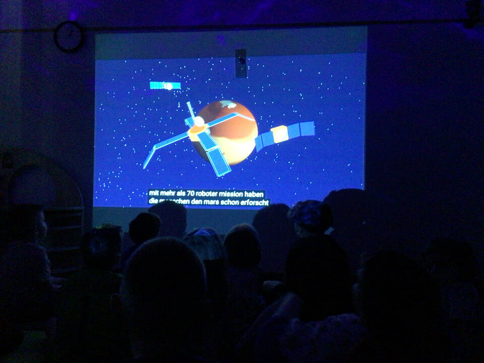 In einem dunklen Raum sieht man ein Bild eines Sateliten.