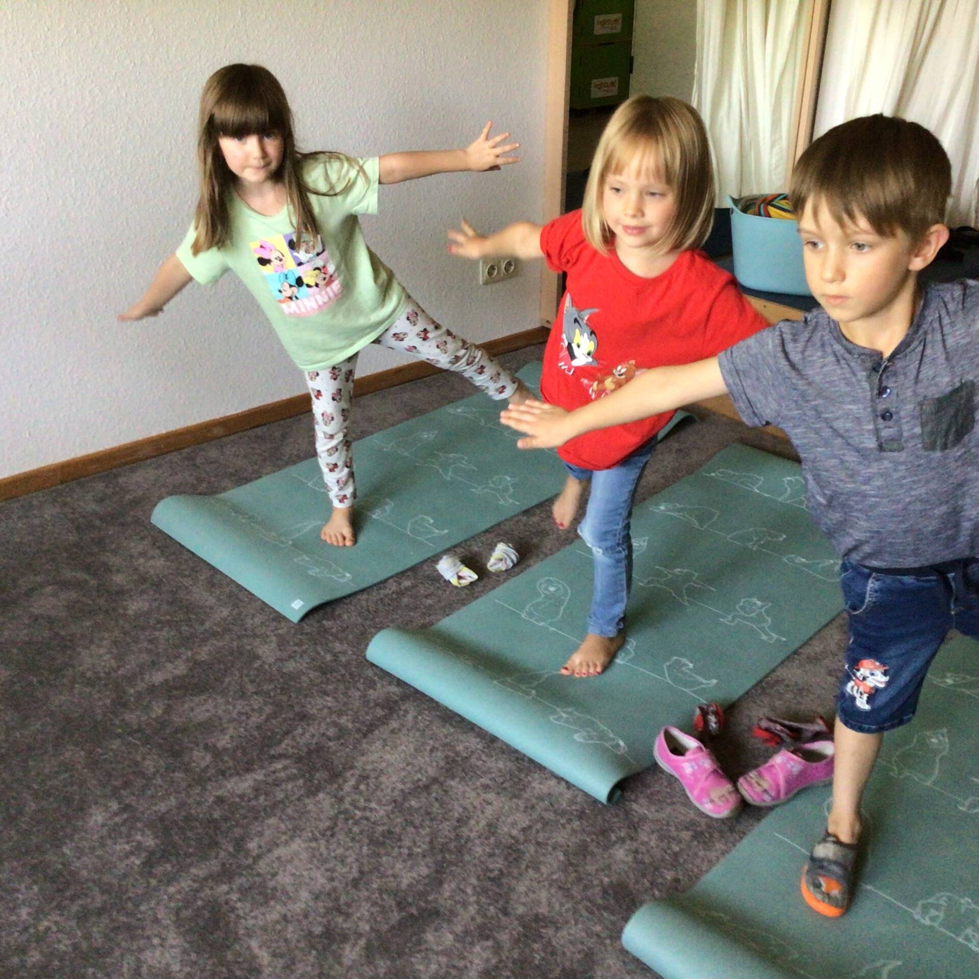Drei Kinder stehen auf einer Matte mit einem Bein, strecken die Arme vom Körper ab.