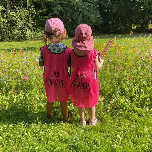 Zwei Kinder mit rosa Kappen und Leibchen auf einer Wiese.