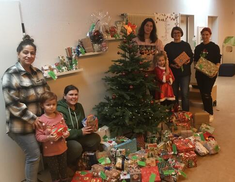 Mehrere jüngere und ältere Personen stehen neben dem Weihnachtsbaum mit Geschenken.