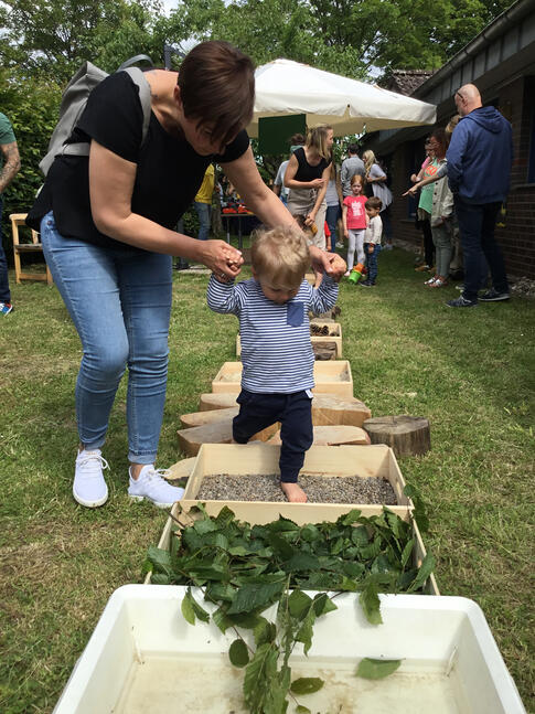 Frau hält Jungen an Händen fest, er läuft durch Kästen mit unterschiedlichem Material.