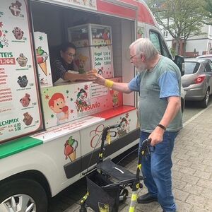 Der Eisverkäufer gibt einem Herrn mit Rollator sein Eis.