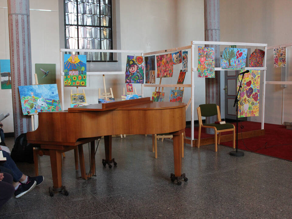 Man sieht einen Klavierflügel und selbstgemalte Bilder in einer Kirche.
