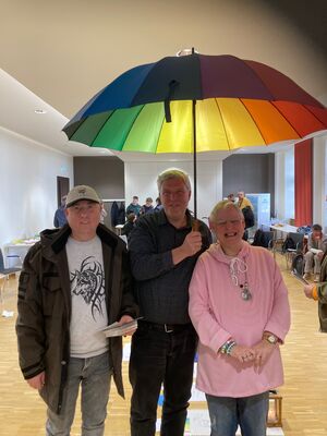 Drei Personen stehen unter einem Regenschirm, mit Regenbogenfarben in einem Raum.