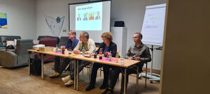 Die Politiker sitzen vor einer Leinwand an Tischen.
