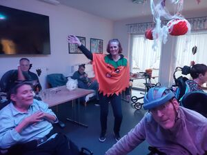 Eine Person alss Kürbis verkleidet steht in der Mitte des Raumes und hebt einen Arm in die Luft.