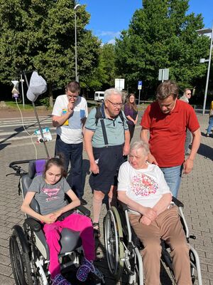 Mehrere Personen stehen in der Sonne, zwei sitzen im Rollstuhl.