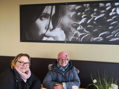 Zwei Personen sitzen innen an einem Tisch, eine Person hat eine rosa Mütze auf.