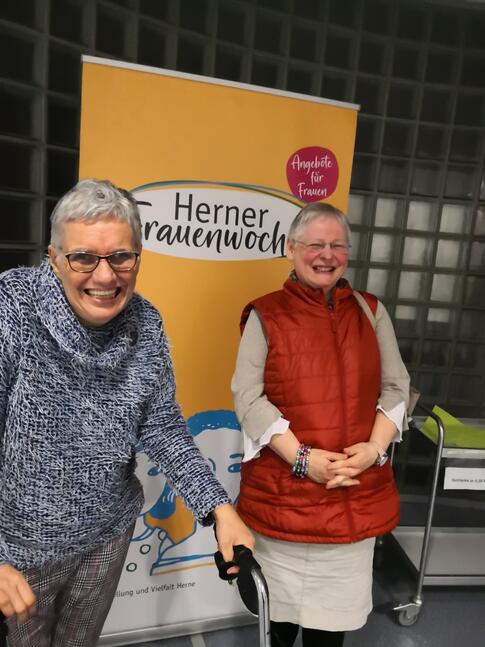 Zwei Frauen lachend vor einem großen Plakat.