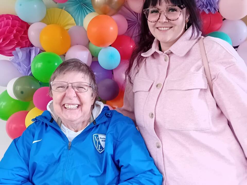 Zwei Personen vor einer Wand mit bunten Luftballons.