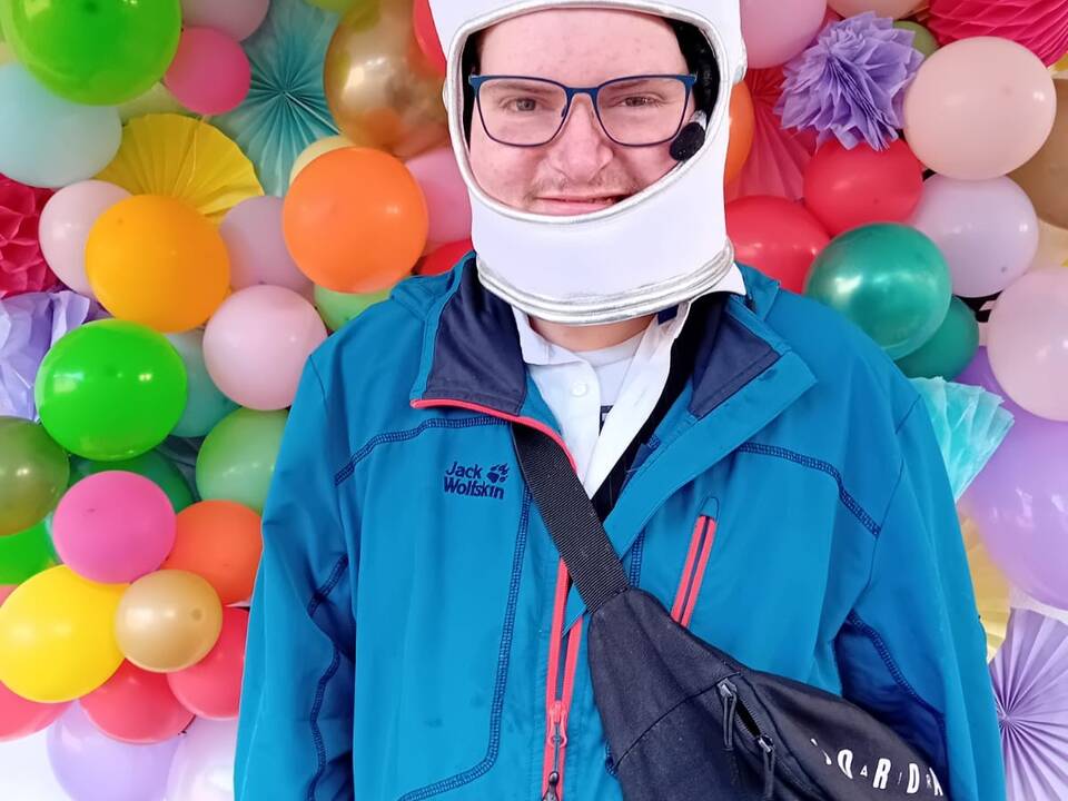 Eine Person mit einer lustigen Kopfbedeckung vor einer Wand mit bunten Luftballons.